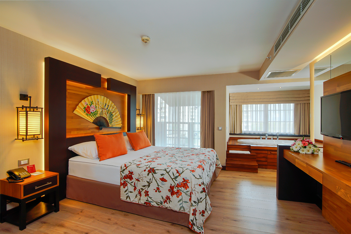 Modern resort bedroom with wooden furnishings, orange accent pillows, decorative fan artwork above the bed, and floor-to-ceiling windows overlooking a balcony.