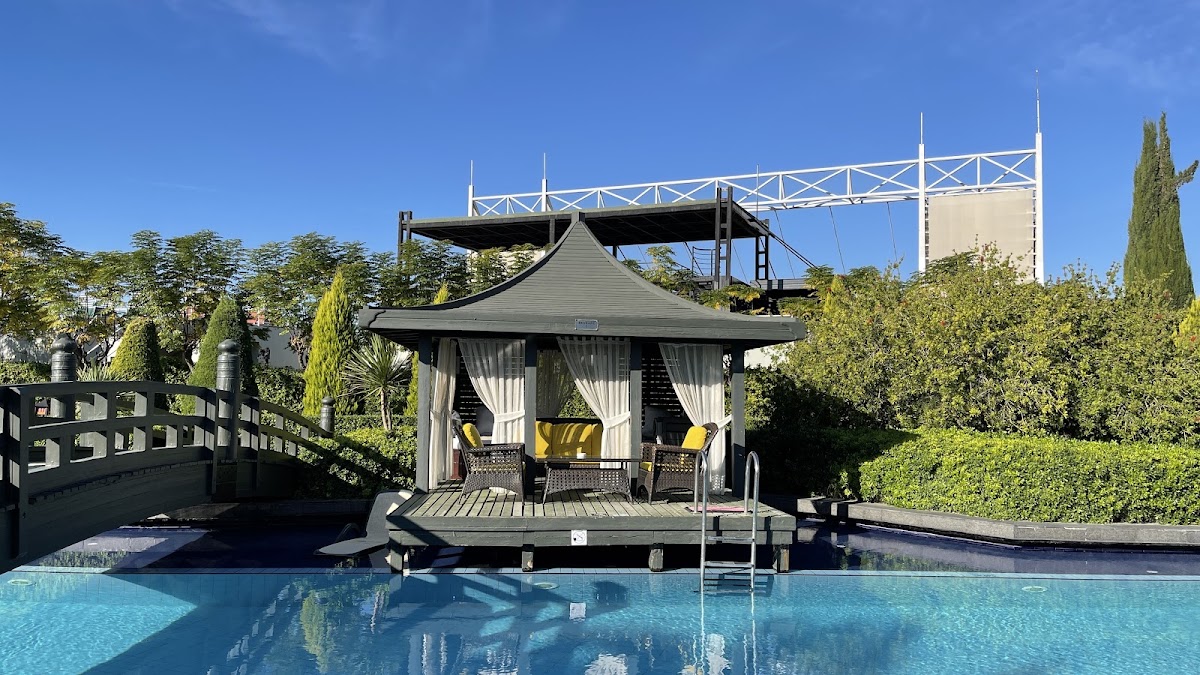 Pagoda-style cabana with yellow chairs sits beside a bright blue pool at Limak Lara De Luxe Hotel & Resort, with a wooden bridge and landscaped grounds visible.