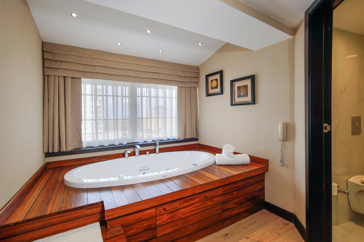 Bathroom with sunken oval bathtub set into rich wood platform, large window with sheer curtains, recessed lighting, and framed botanical prints on beige walls.