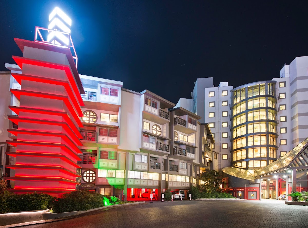 Night view of Limak Lara De Luxe Hotel & Resort showing illuminated buildings with red neon signage and lit windows around a paved courtyard.