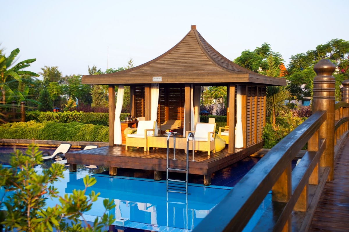 Wooden cabana with pointed roof sits on a deck beside a bright blue pool, furnished with white cushioned chairs and surrounded by lush tropical gardens.