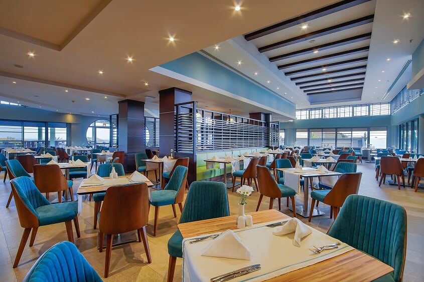Spacious dining room with wooden tables, teal and cognac chairs, striped ceiling beams, and floor-to-ceiling windows overlooking the coast.