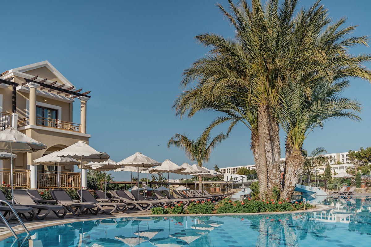Resort pool area with lounge chairs, white umbrellas, and tall palm trees under clear blue sky at Lindos Imperial Resort & Spa.