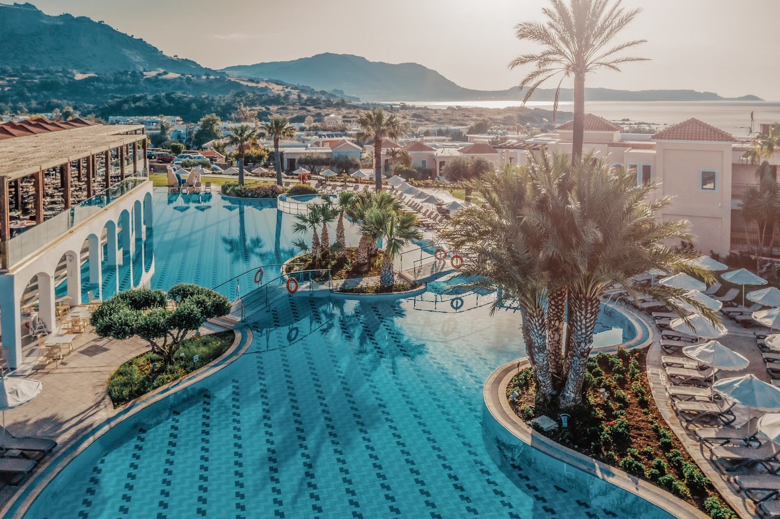 Large turquoise lap pool with lane markers at Lindos Imperial Resort, palm trees, loungers, and coastal mountains beyond.