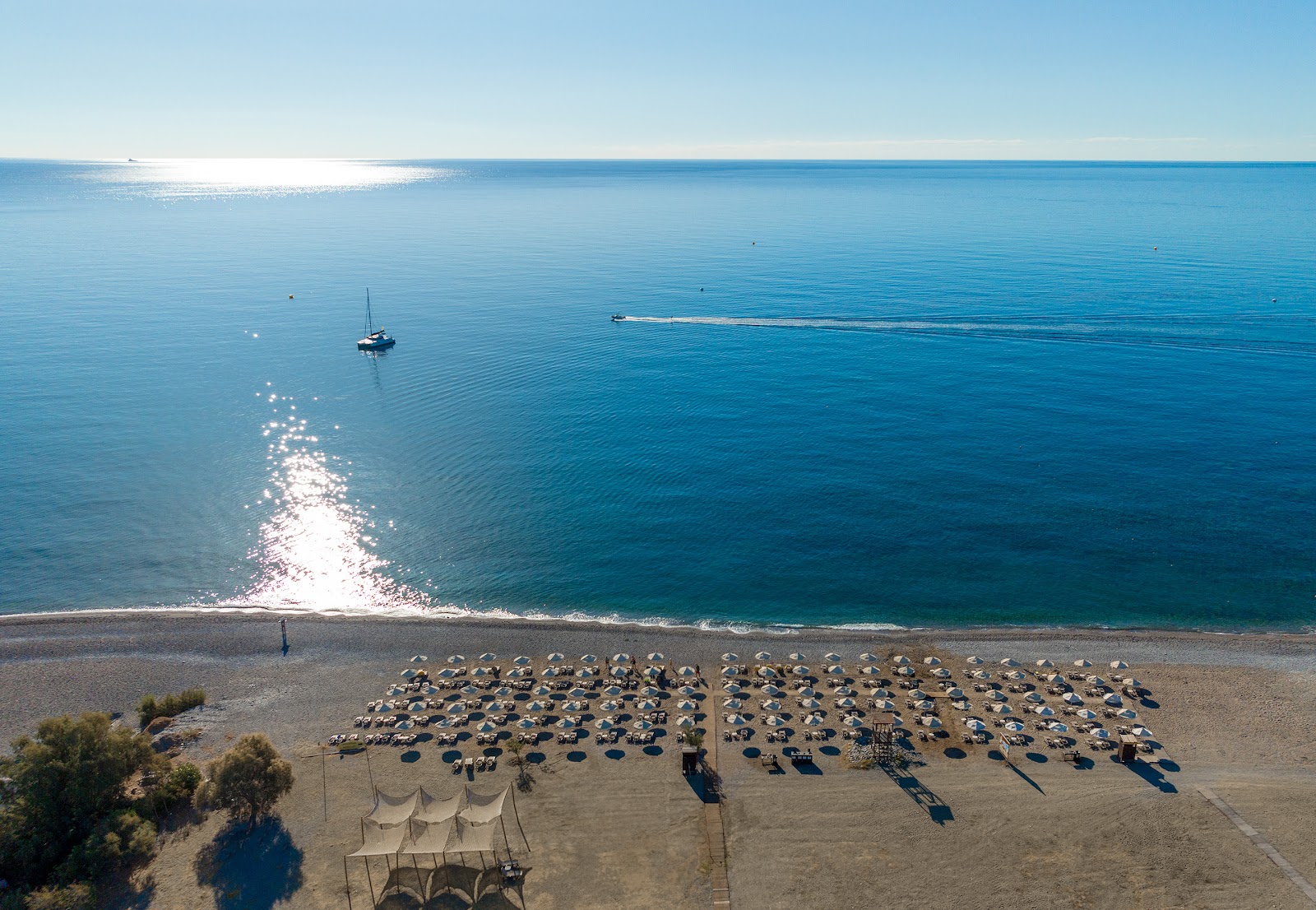 Organized rows of blue and white umbrellas and loungers fill the sandy beach at Lindos Imperial Resort & Spa, with calm turquoise water and moored sailboats beyond.