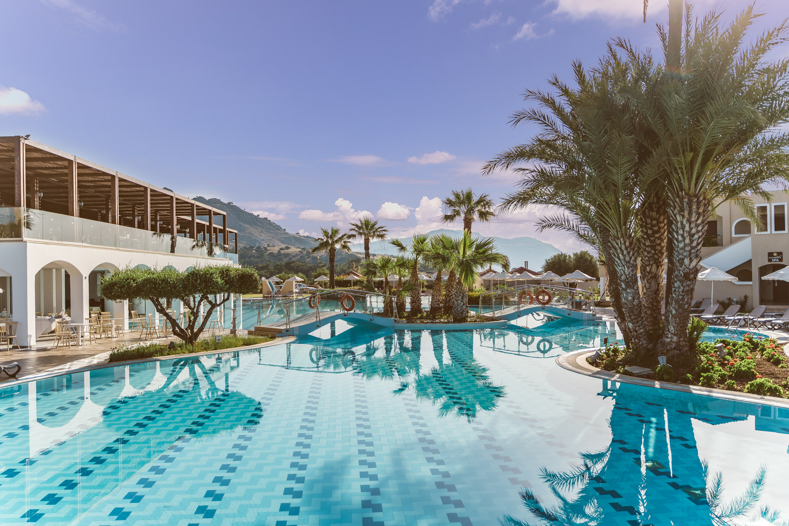 Large resort pool with geometric sections, palm trees, and white buildings with arched details against mountains and sea views.
