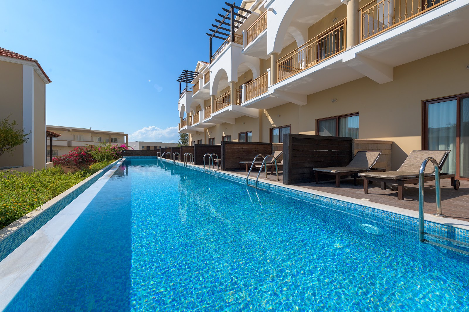 Shaded terrace with loungers overlooks Lindos Imperial Resort & Spa's pool, beach, and sea under clear blue sky.