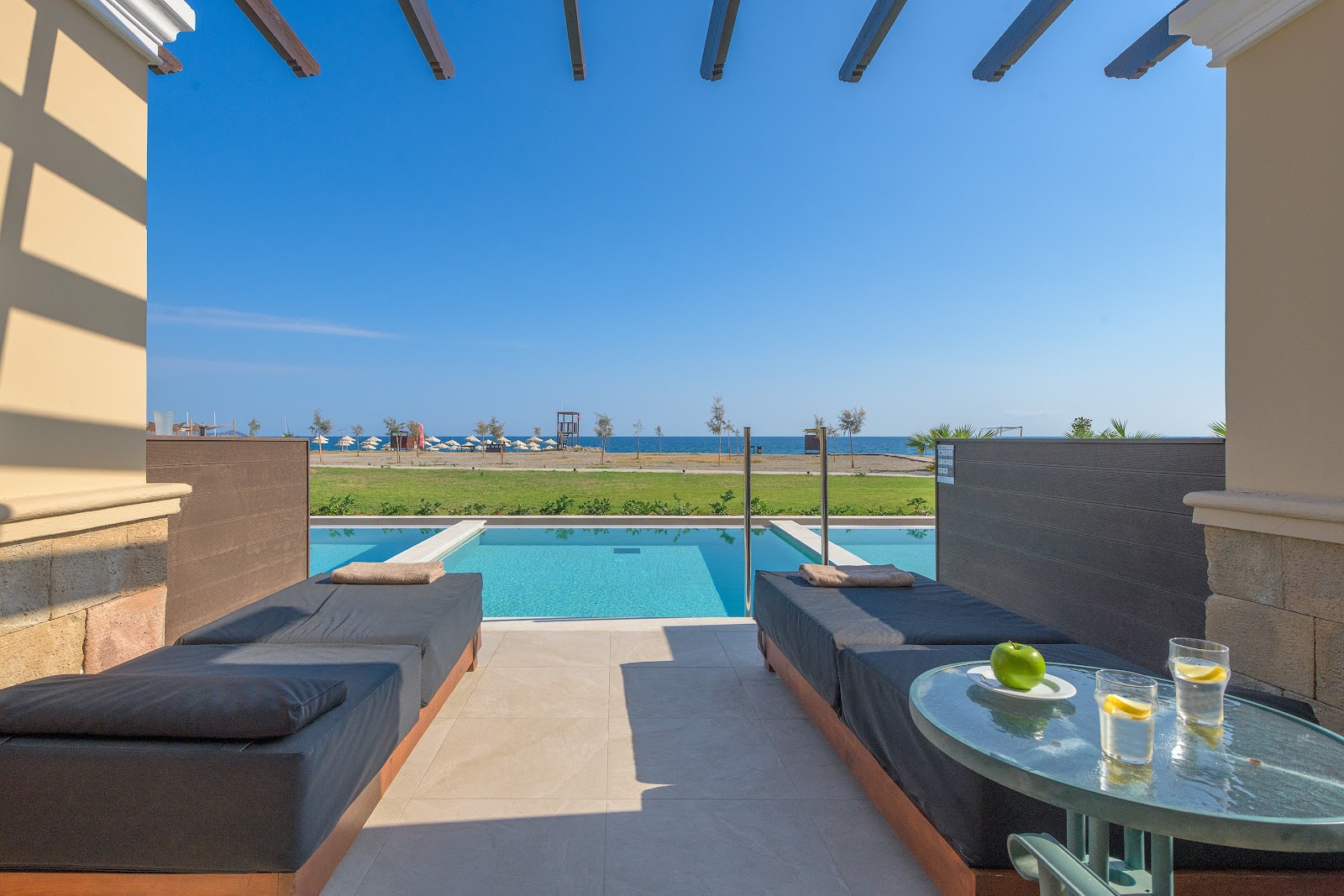 Long swimming pool with lounge chairs and dark privacy screens on wooden deck beside multi-story white resort buildings with arched balconies.