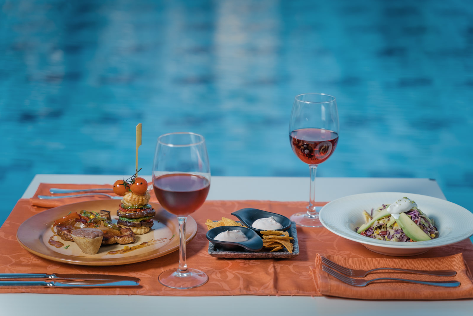 Poolside dining table with two plated dishes, red wine glasses, and an orange tablecloth overlooking the pool at Lindos Imperial Resort & Spa.