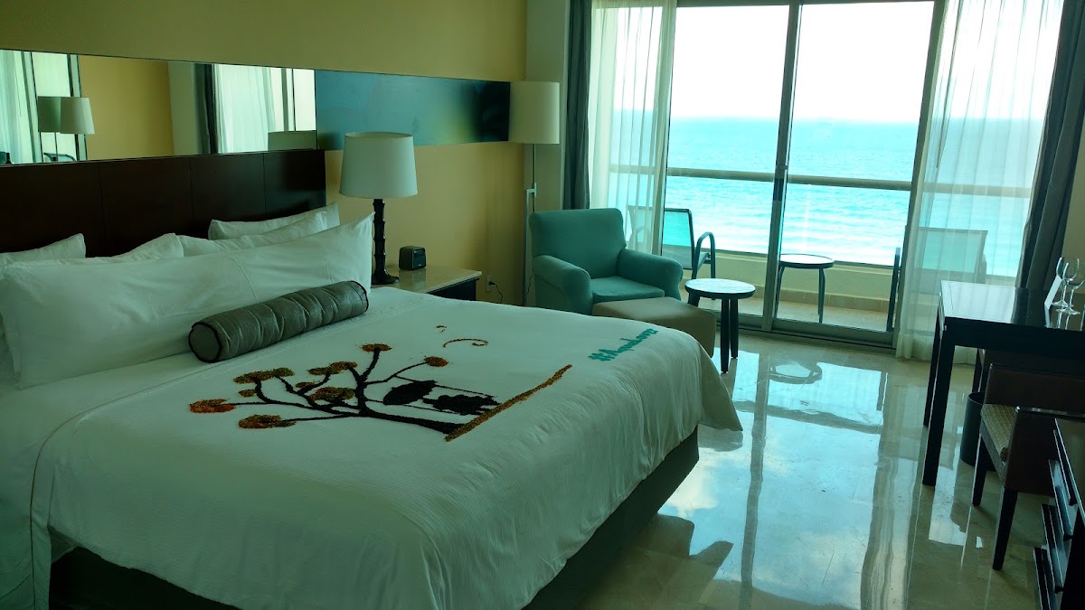 Oceanfront resort bedroom with white bed, teal seating area, and turquoise sea view through floor-to-ceiling windows.