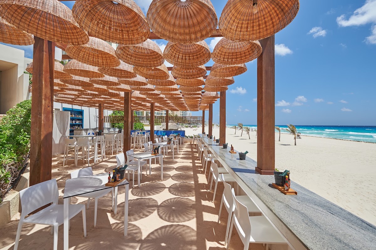 Woven basket shade structures cover a beachfront dining area with white chairs at Live Aqua Beach Resort Cancún, overlooking turquoise Caribbean waters.