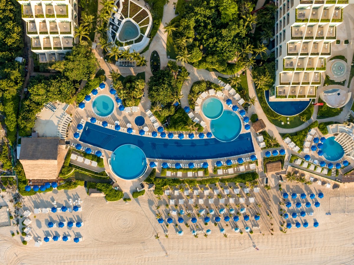 Overhead view of Live Aqua Beach Resort Cancún with multiple turquoise pools, blue umbrellas, loungers on sandy beach, and lush tropical greenery surrounding the grounds.
