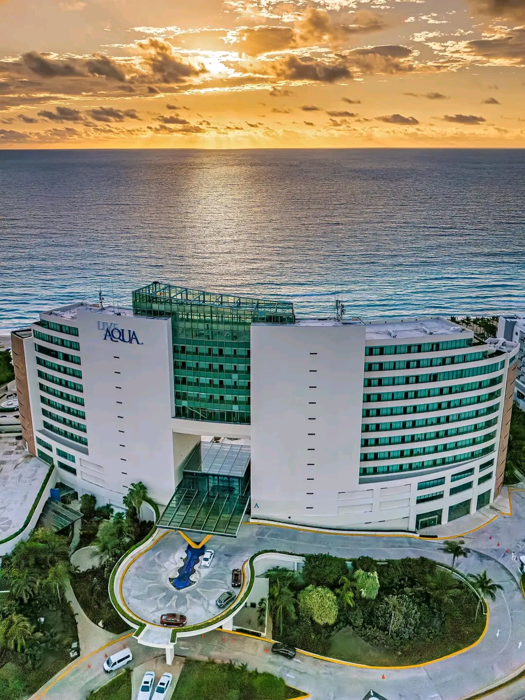Live Aqua Beach Resort overlooks the ocean at sunset, with curved white and green towers flanking a central entrance and landscaped grounds below.