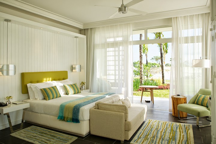 Bright bedroom with chartreuse headboard, striped pillows, and sheer curtains framing glass doors opening to palm trees and grounds at Long Beach Mauritius.