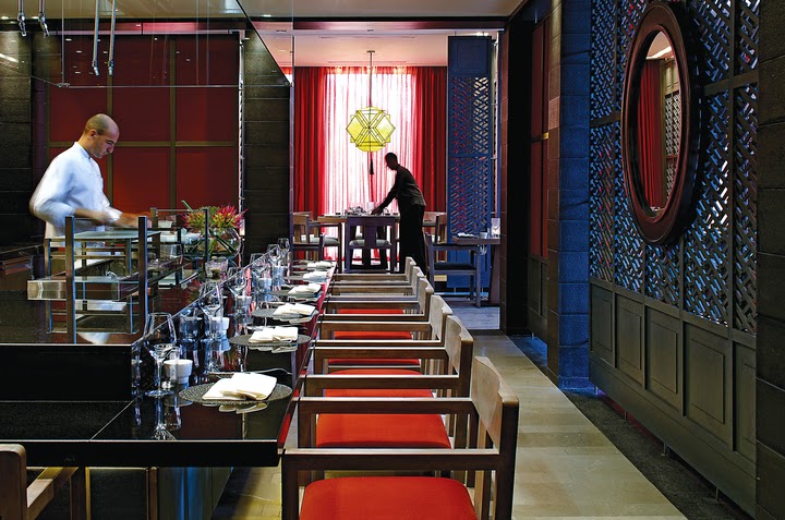 Restaurant dining room with red cushioned chairs at tables set with glassware, staff preparing service, and bold blue and red decor throughout.