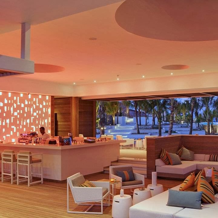 Modern resort lounge with coral ceiling, bar seating, and pink upholstered furniture overlooking palm trees and beach at sunset.