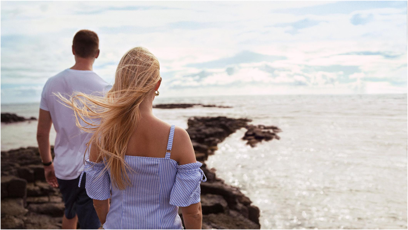 Couple standing on rocky shoreline facing calm ocean waters under partly cloudy sky.