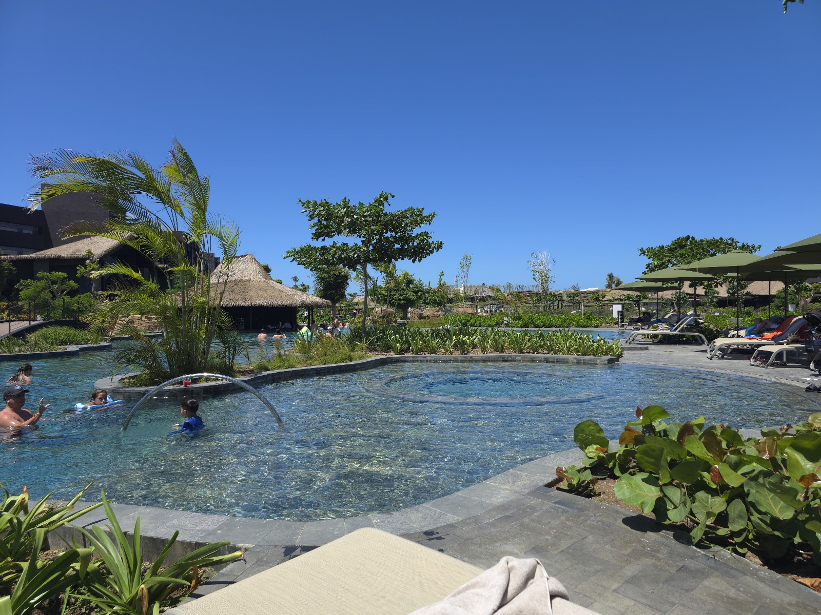 Resort pool with guests swimming, surrounded by palm trees, loungers with green umbrellas, and a thatched-roof structure in the background under clear blue sky.