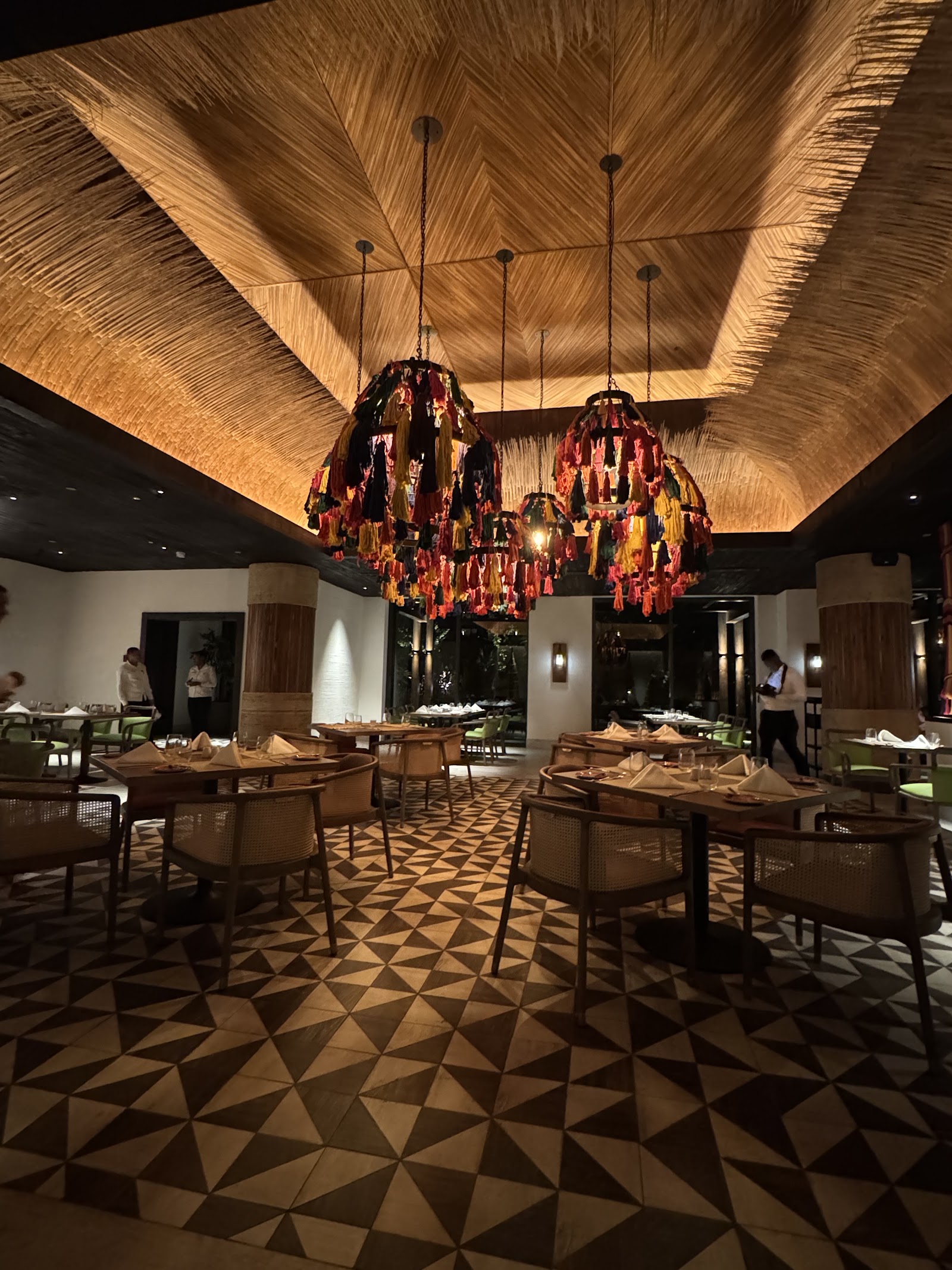 Upscale dining room with woven ceiling, colorful tassel chandeliers, geometric floor pattern, and tables set for dinner service.