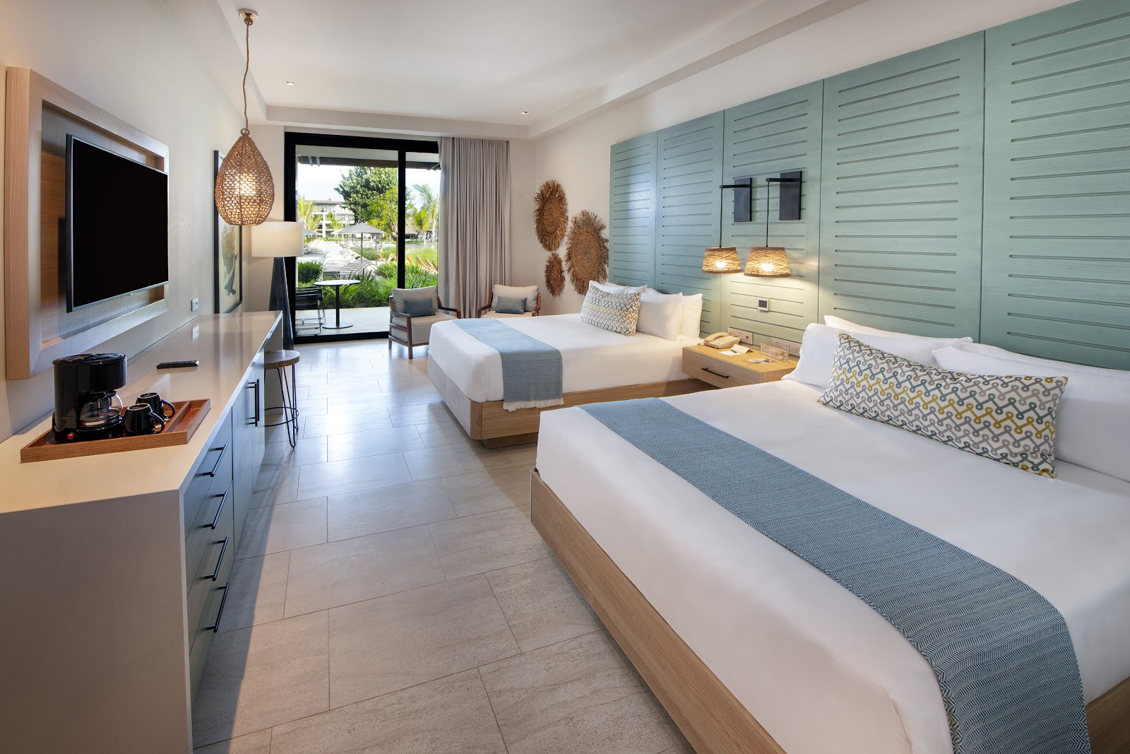 Spacious resort room with two queen beds, light wood furnishings, turquoise closets, and a balcony overlooking tropical grounds.