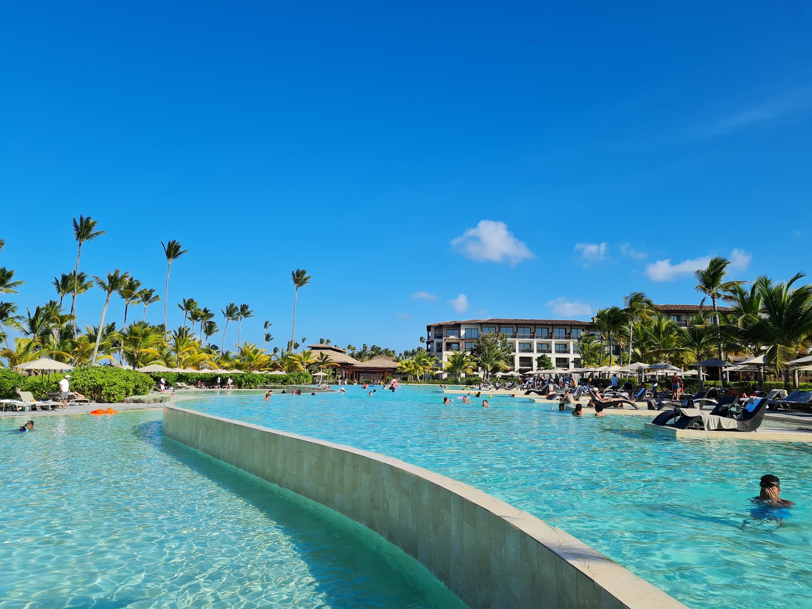 Large lagoon-style pool with concrete divider, guests swimming, palm trees, white umbrellas, and multi-story resort building under clear blue sky.
