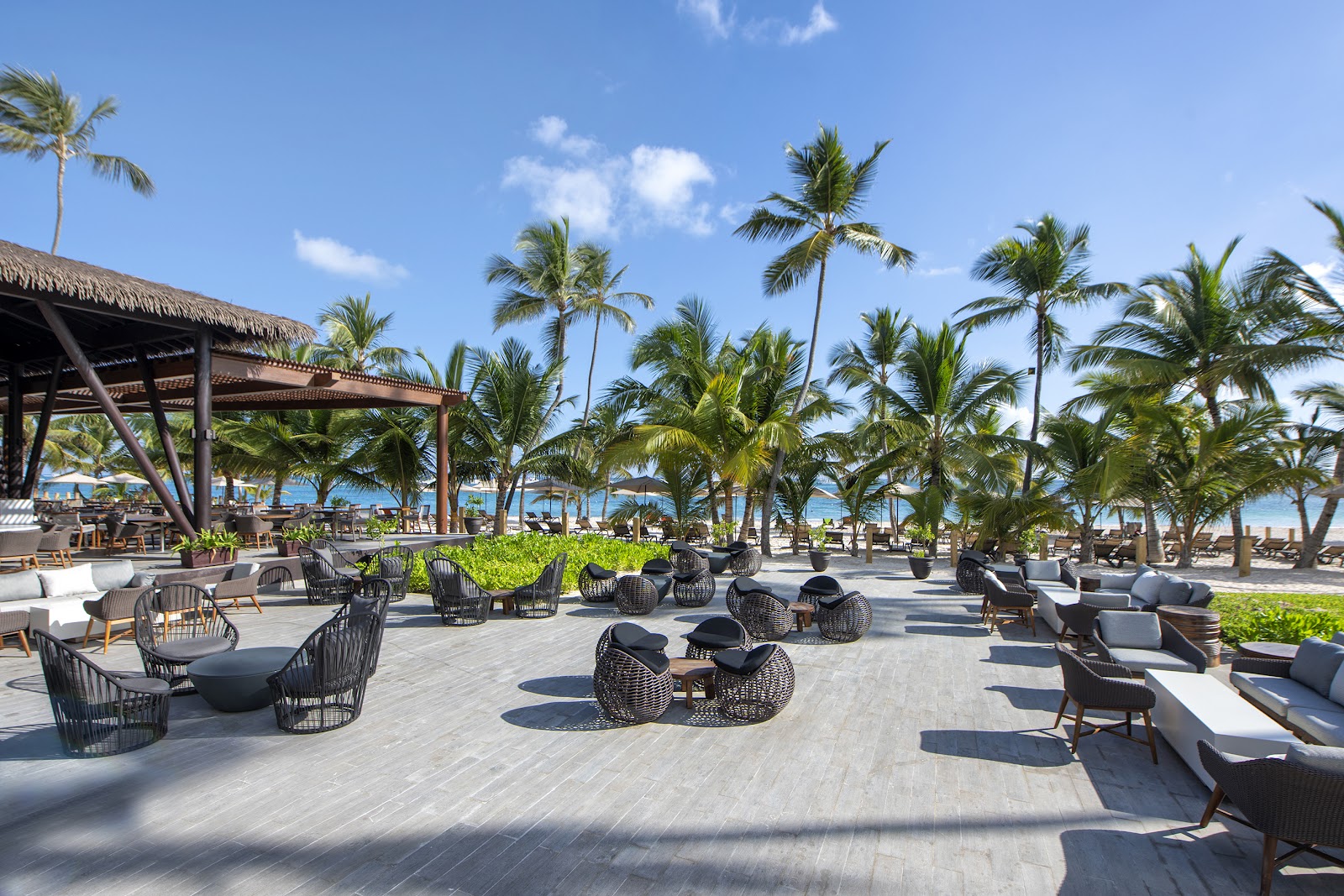 Beachfront lounge area with black wicker chairs and white cushions scattered across a paved terrace, palm trees, and ocean view in the background.