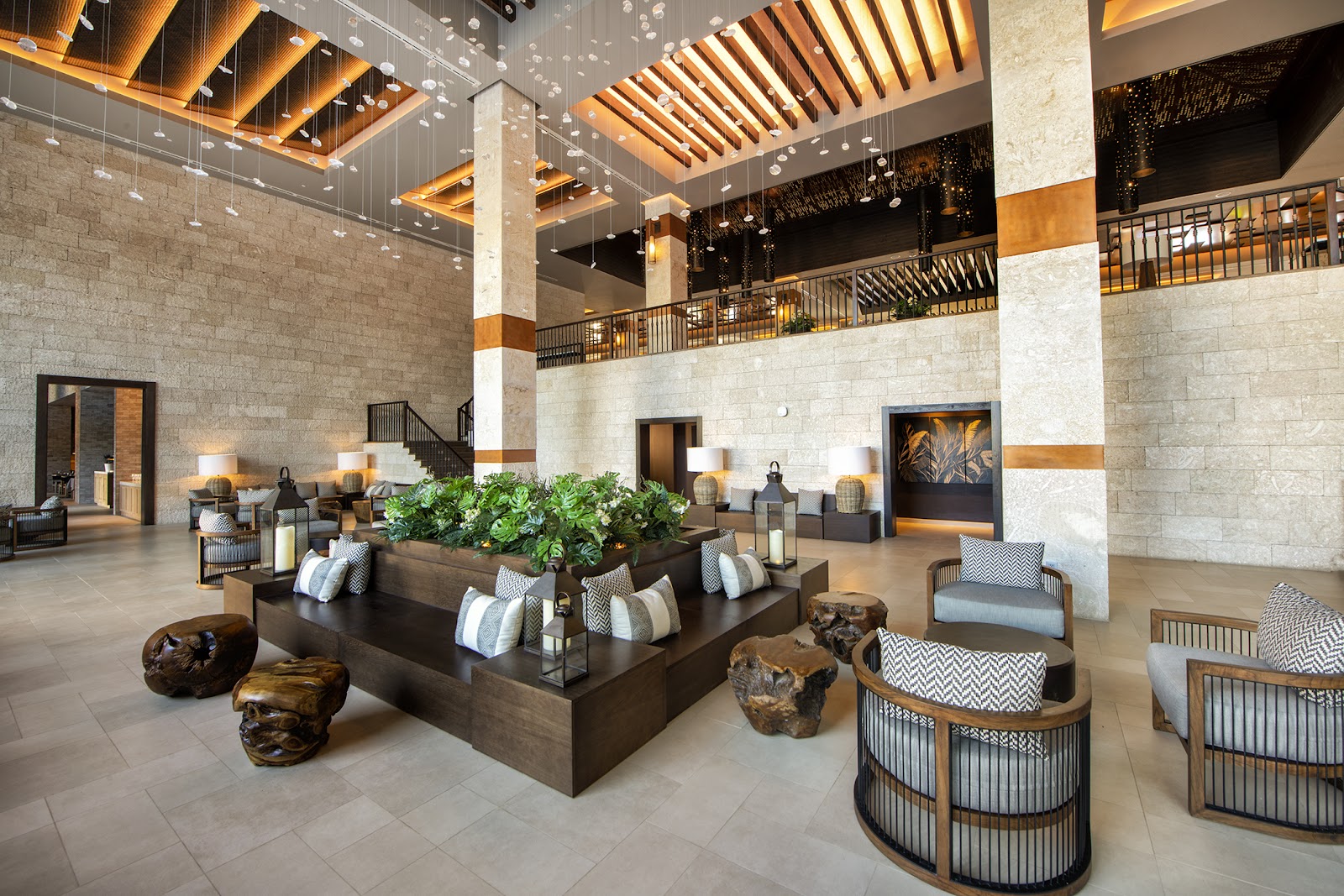 Spacious resort lobby with stone walls, hanging pendant lights, wooden beams overhead, and a central planter surrounded by seating areas.