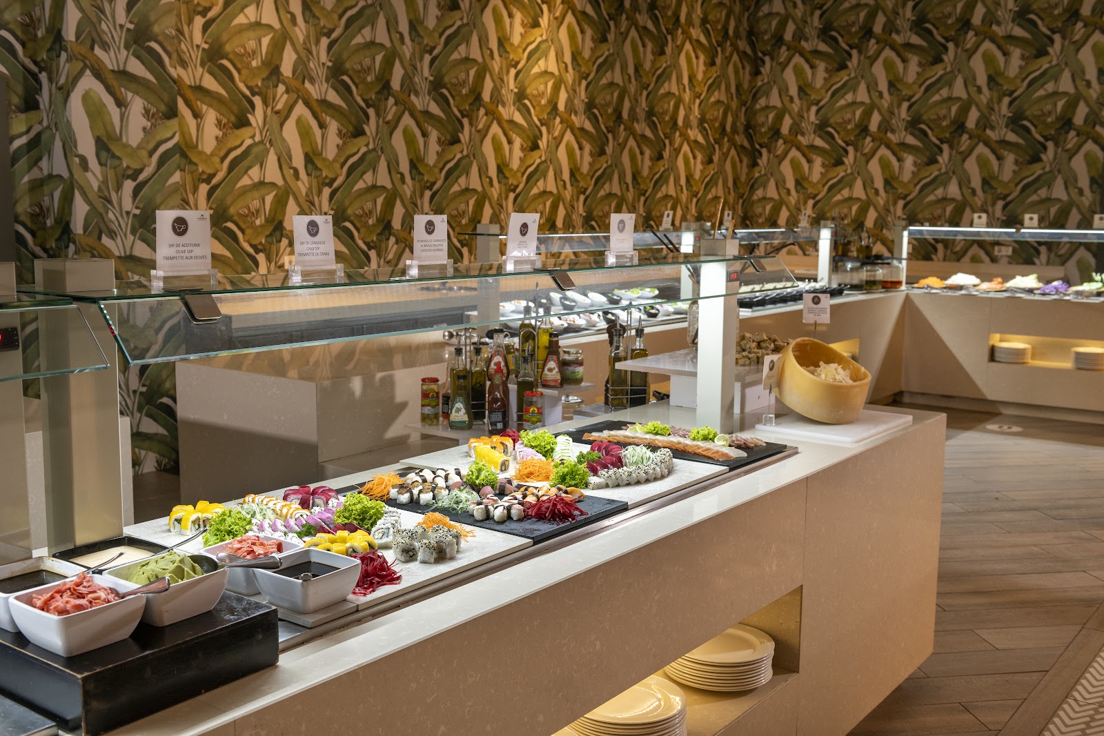 Buffet line at Lopesan Costa Bavaro Resort displays colorful salads, cured meats, sushi, and garnishes under heat lamps with labeled food stations and a leafy botanical wallpaper backdrop.