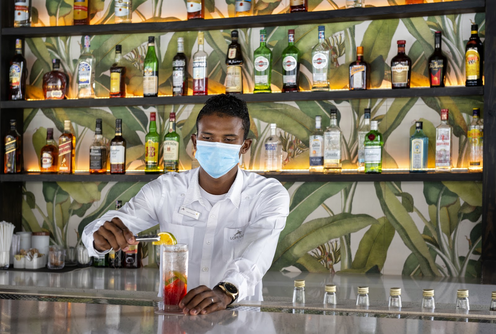 Bartender wearing mask prepares a cocktail at a backlit bar with tropical leaf wallpaper and shelves of spirits.