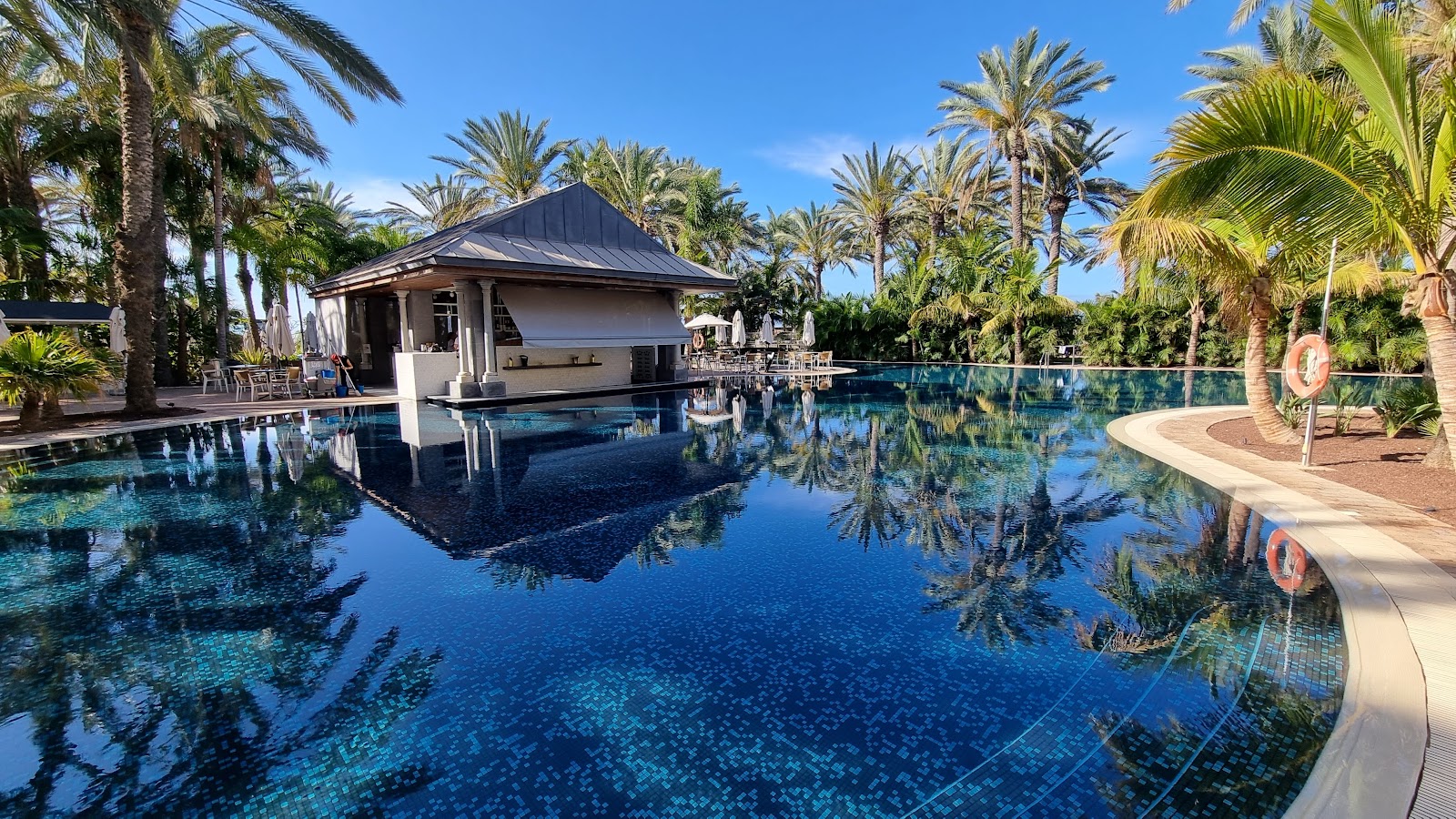 Large curved pool with blue tile and a dark-roofed poolside bar surrounded by tall palm trees under clear sky at Lopesan Costa Meloneras.