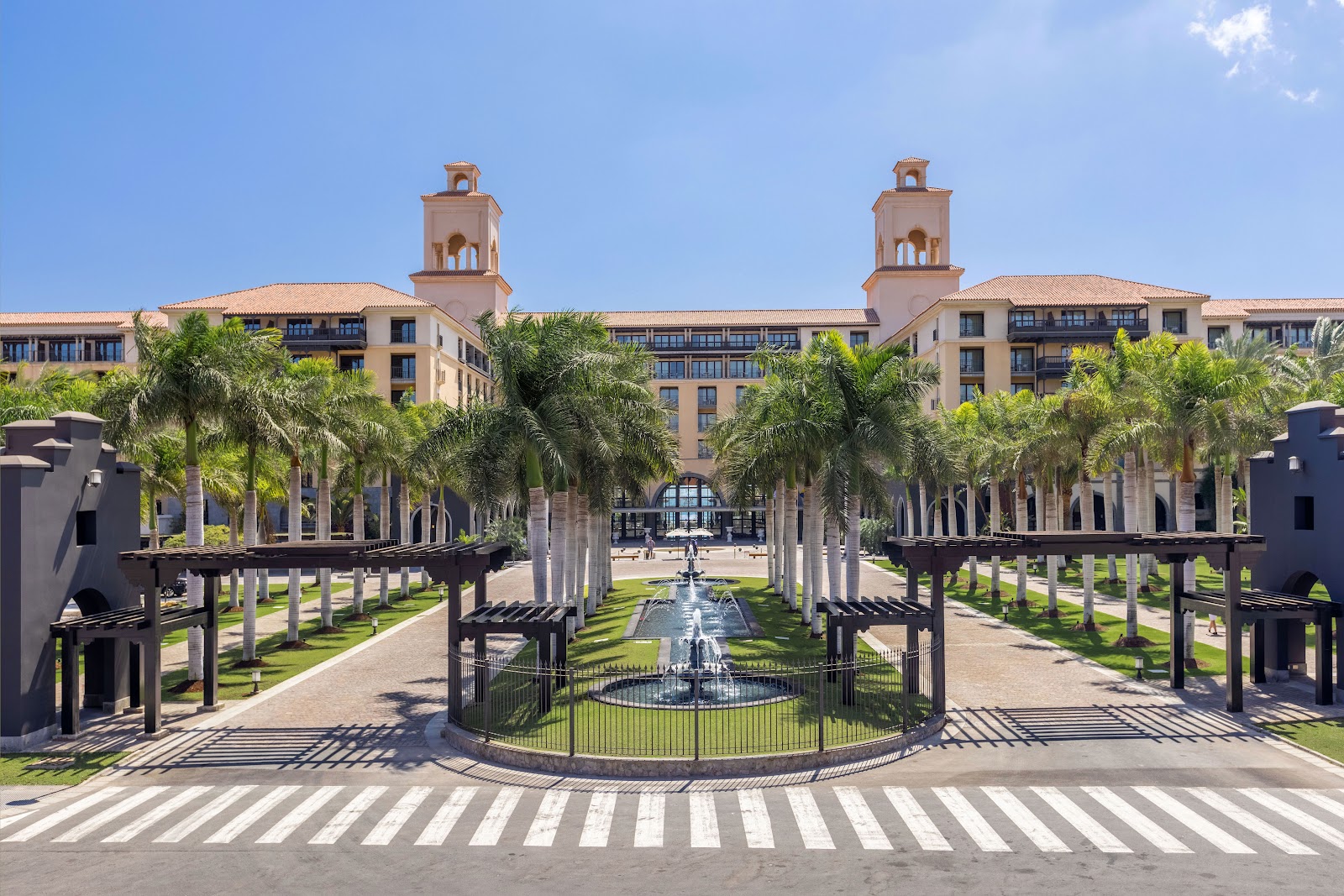 Central courtyard with fountain, palm trees, and Spanish colonial architecture with two bell towers under clear sky.