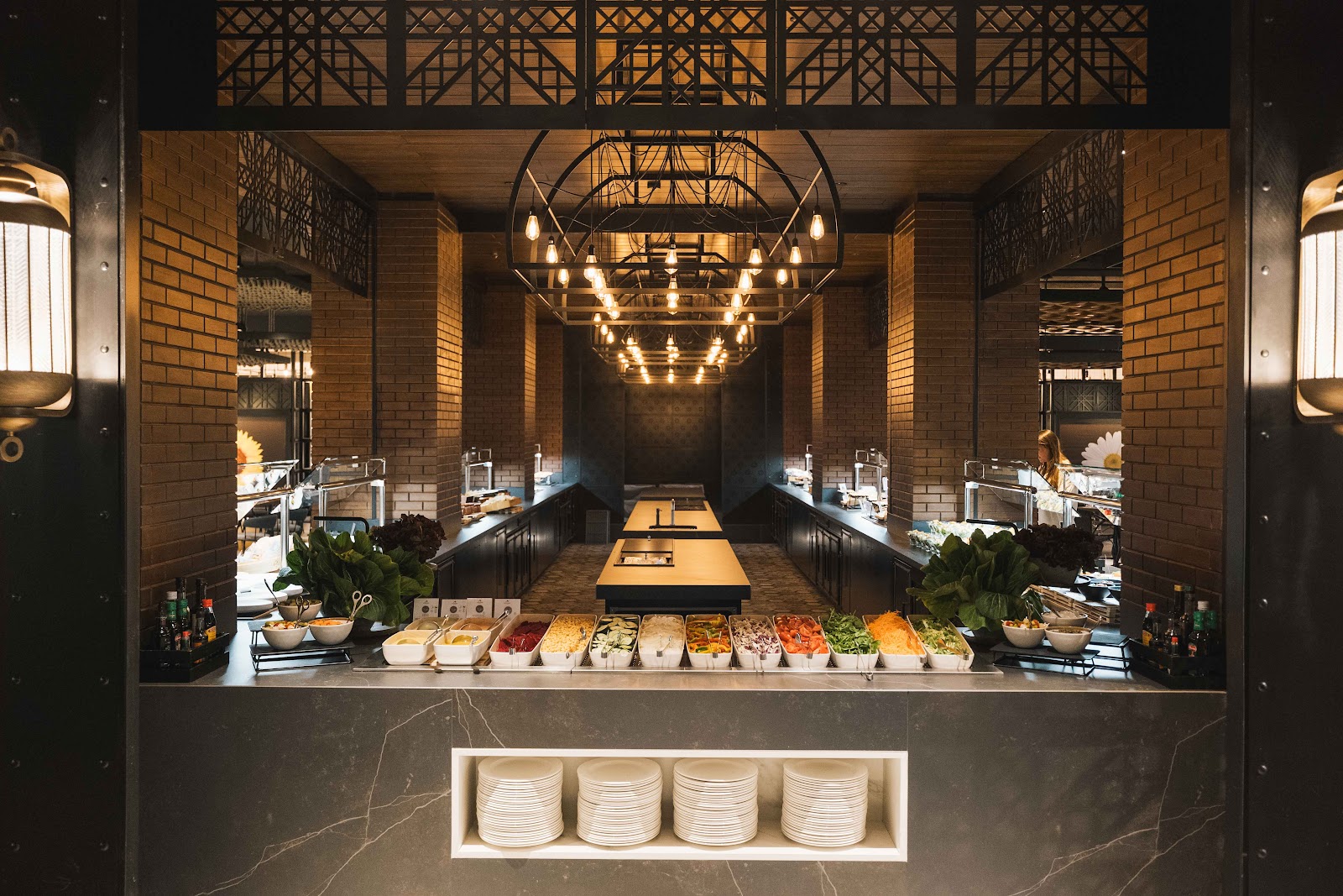 Modern resort buffet station with brick walls, hanging geometric pendant lights, and rows of fresh food containers flanked by bathroom vanities.