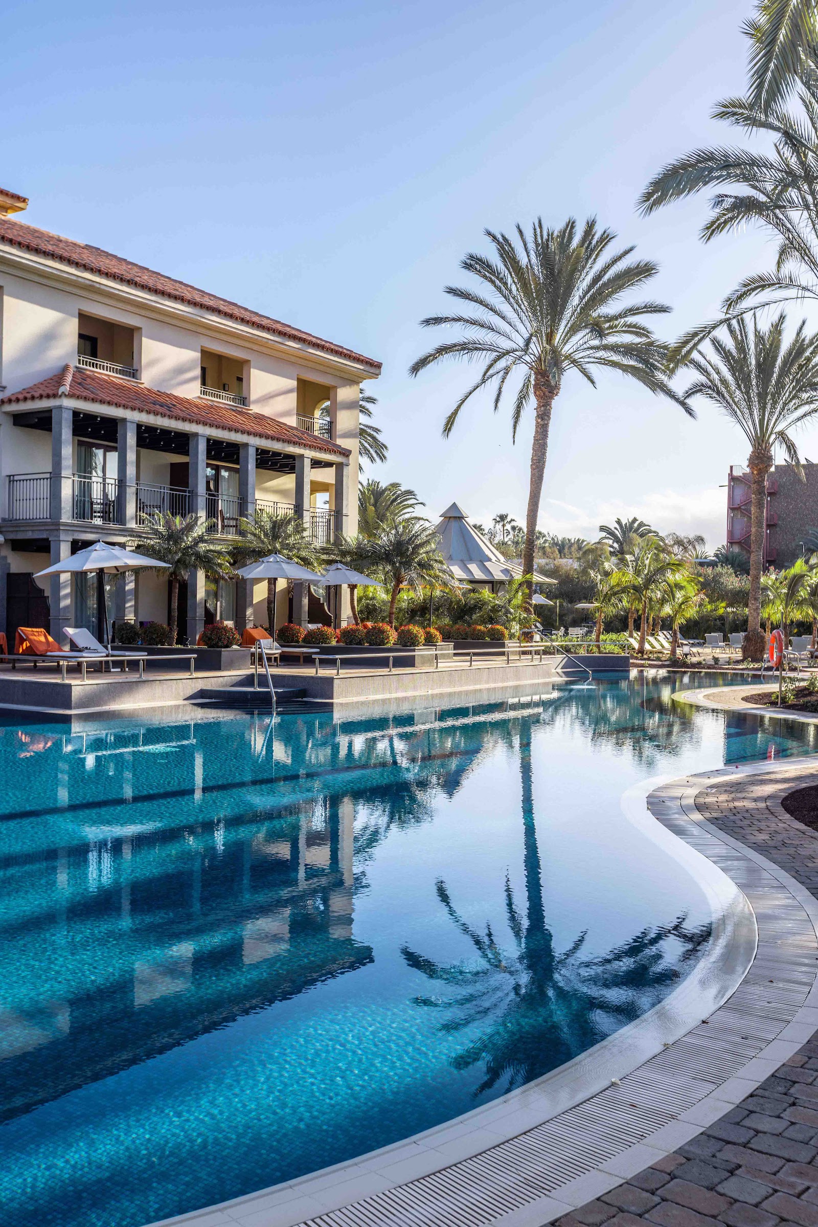 Narrow lap pool lined with loungers and palm trees, flanked by beige resort buildings with balconies and a lighthouse visible in the distance.