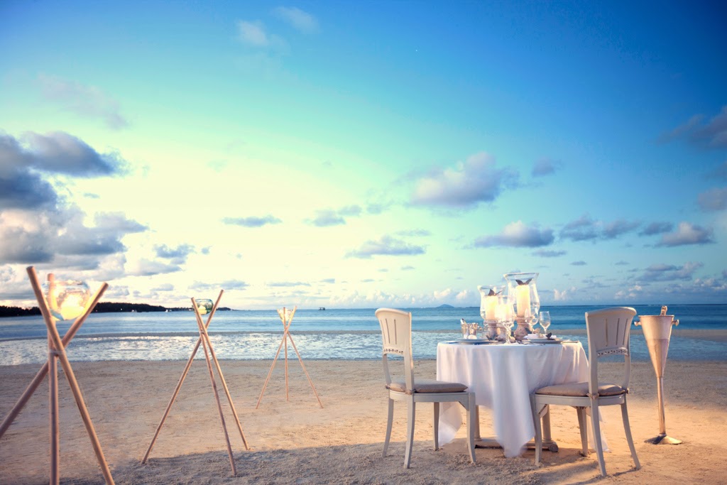 Beach dining setup at LUX* Belle Mare with a white-clothed table, chairs, and torches on sand facing the ocean at dusk.