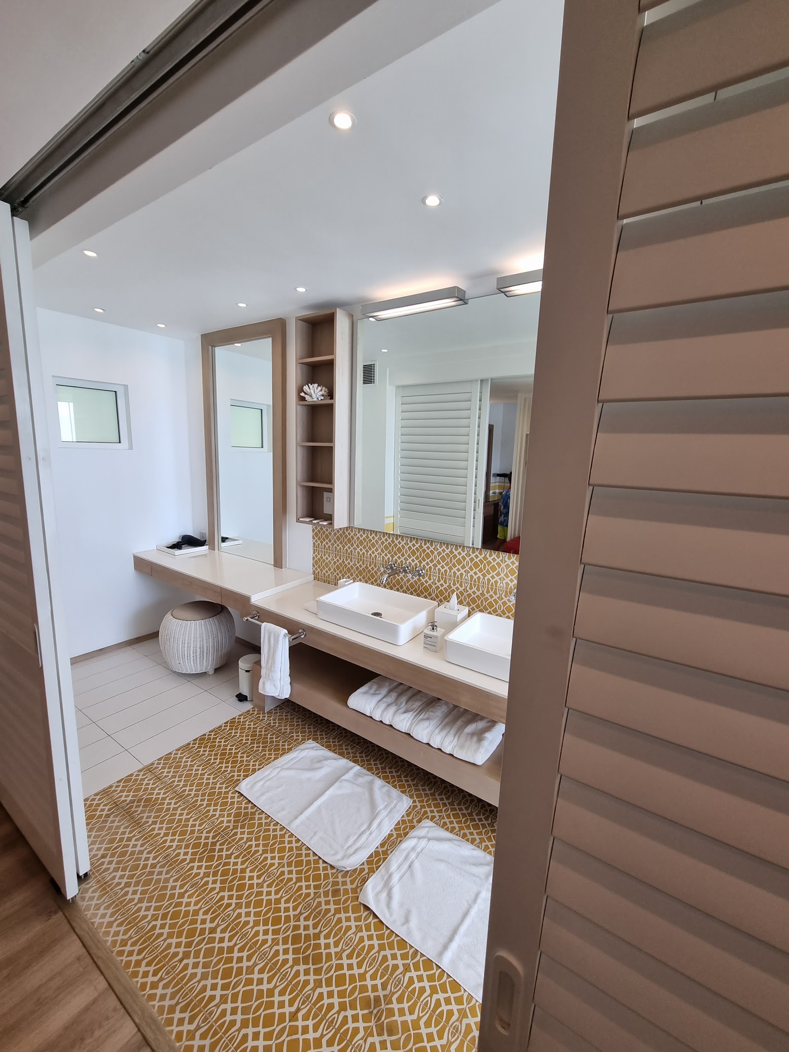 Modern resort bathroom with dual vanities, large mirror, gold geometric floor tiles, white towels, and wooden accents at LUX Grand Gaube.