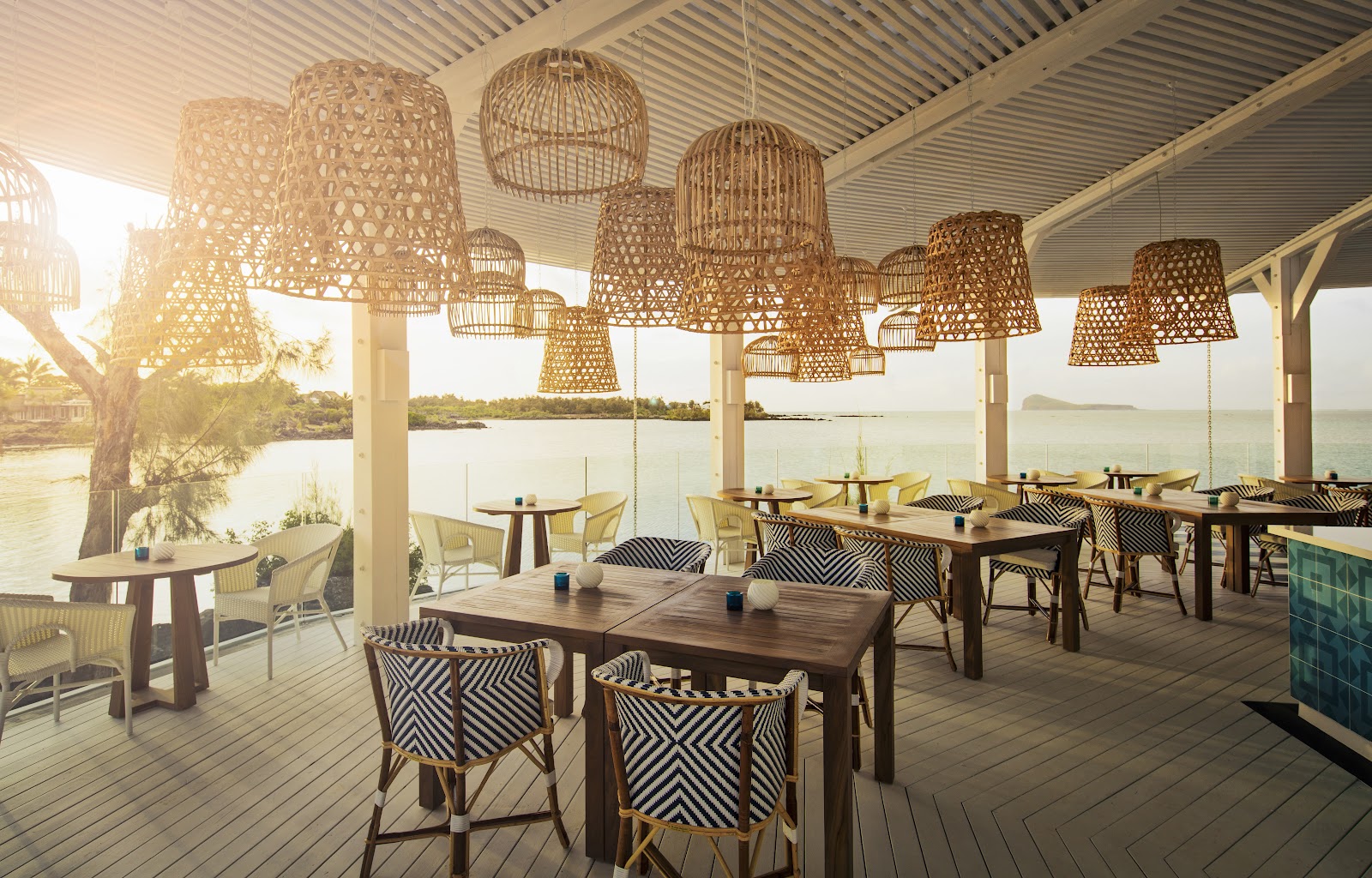Waterfront dining pavilion with woven pendant lights, wooden tables, and striped chairs overlooking calm lagoon and distant islands.
