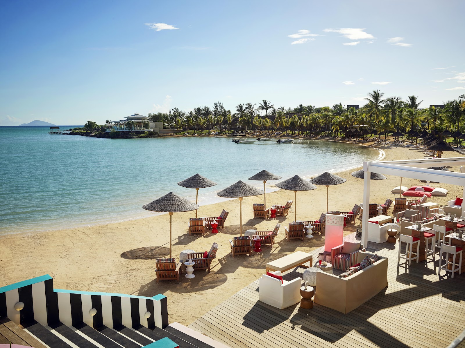 Beach club with thatched umbrellas, wooden loungers, and dining area overlooking turquoise lagoon with palm trees and distant island.