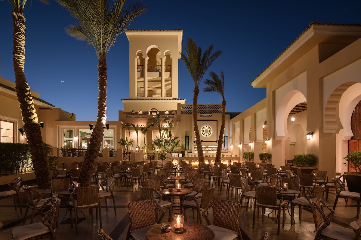 Illuminated courtyard at dusk with woven chairs arranged around tables, palm trees, and a towering Moorish-style building with arched colonnade against a deep blue twilight sky.