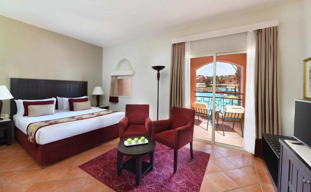 Spacious guest room with king bed, red armchairs, and balcony overlooking the resort pool and Red Sea coastline.