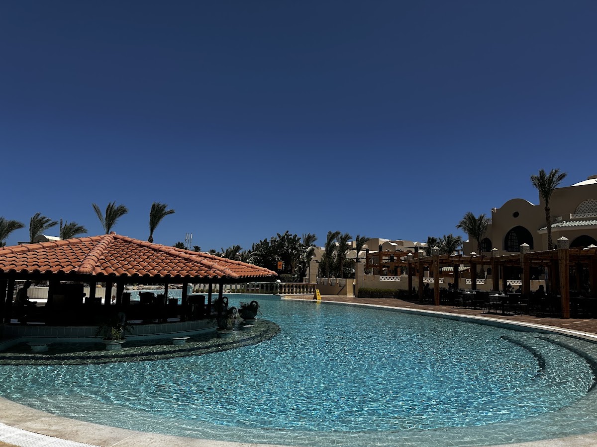 Resort pool with tiled red roof pavilion, palm trees, and tan-colored buildings under clear blue sky.