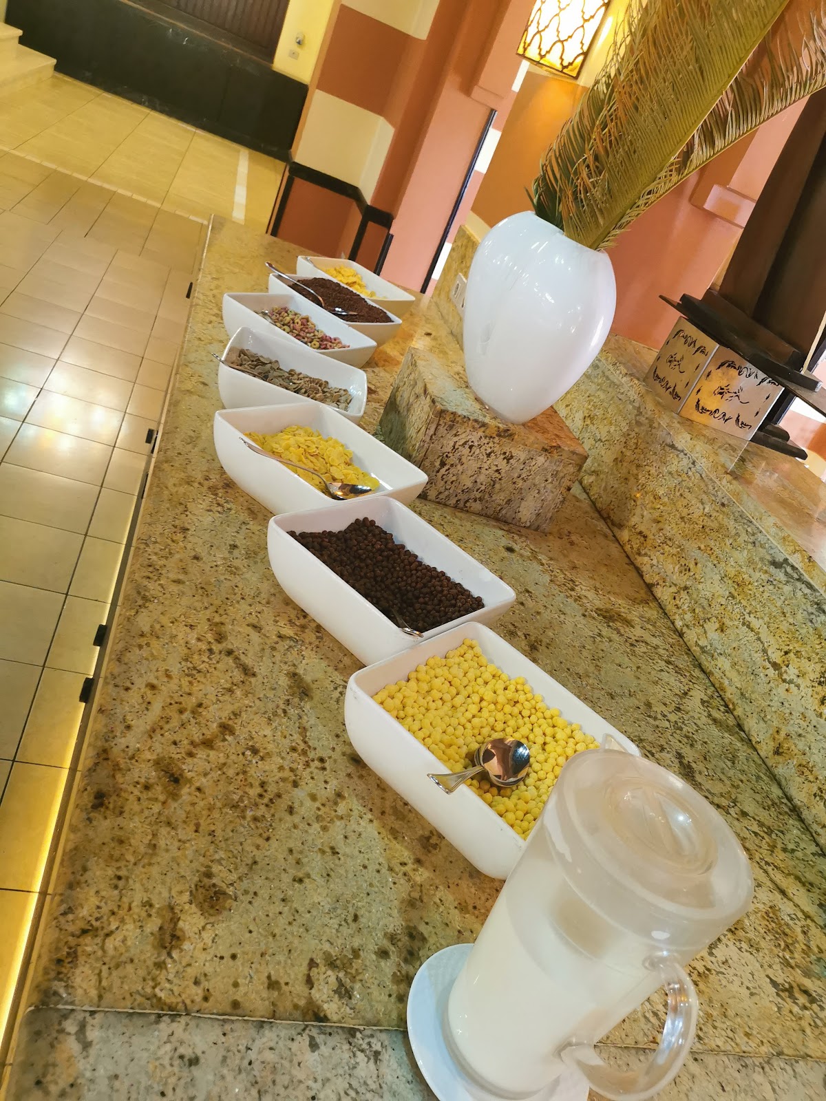 Buffet line with white bowls containing corn, beans, grains, and other ingredients on a granite counter in an orange and coral dining area.