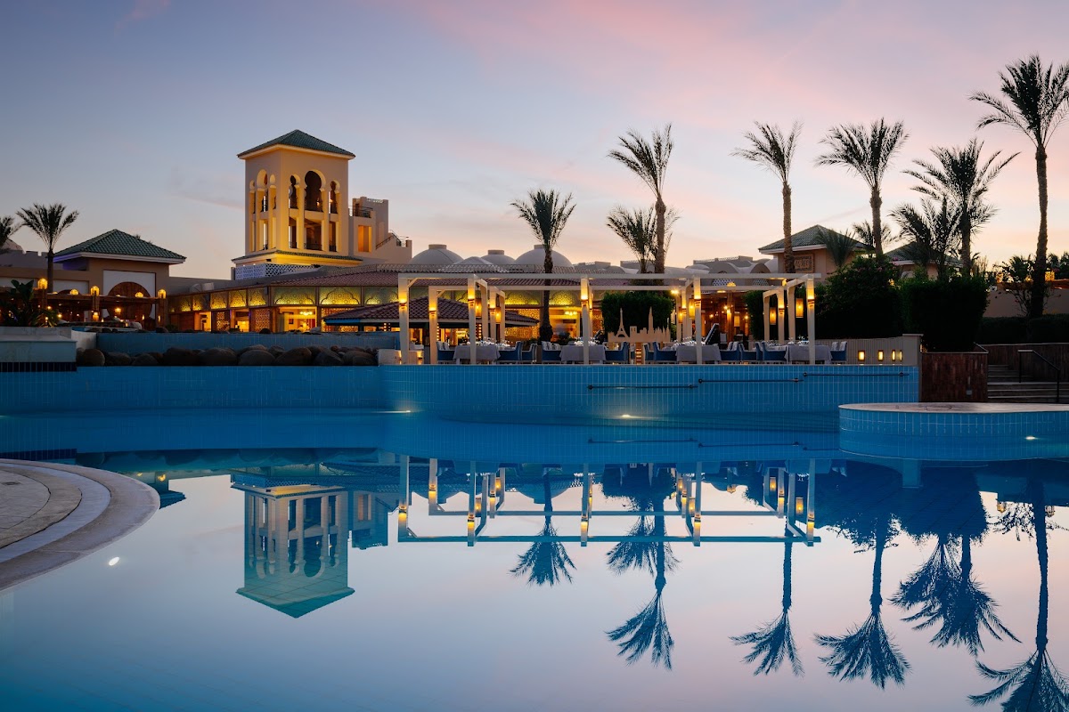 Resort pool at twilight reflects palm trees and illuminated buildings with a yellow tower at dusk.