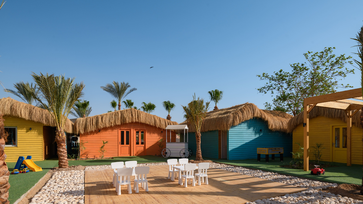 Colorful thatched-roof bungalows in yellow, orange, and blue surround a central courtyard with white dining furniture and palm trees under clear sky.