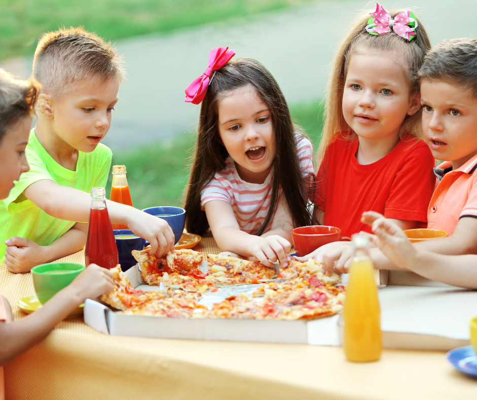Children sharing pizza and drinks at an outdoor table with green lawn in the background.