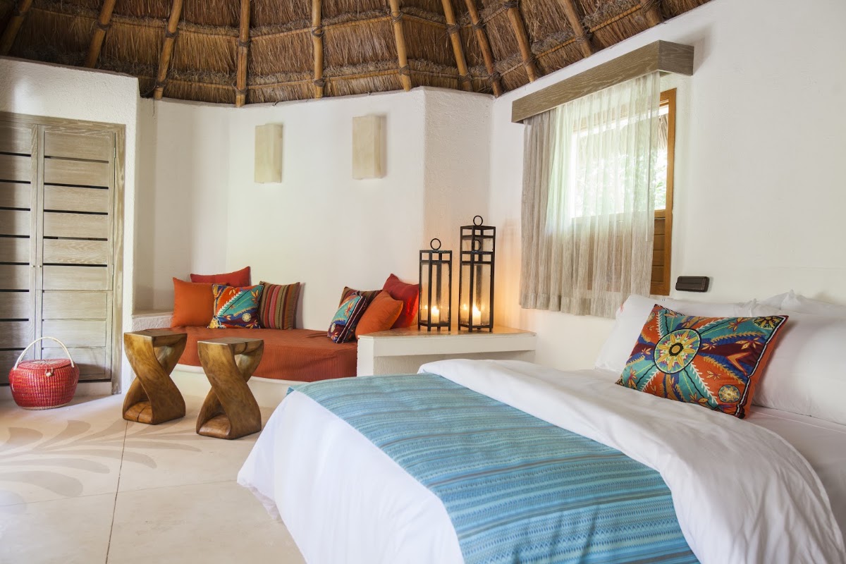 Resort room with thatched roof, queen bed with blue runner, orange seating area, colorful pillows, and wooden side tables.