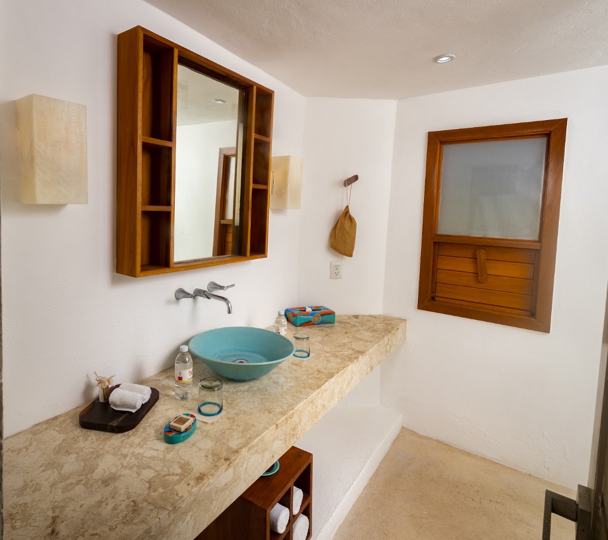 Resort bathroom with turquoise vessel sink, travertine countertop, wooden mirror cabinet, and window overlooking the grounds.