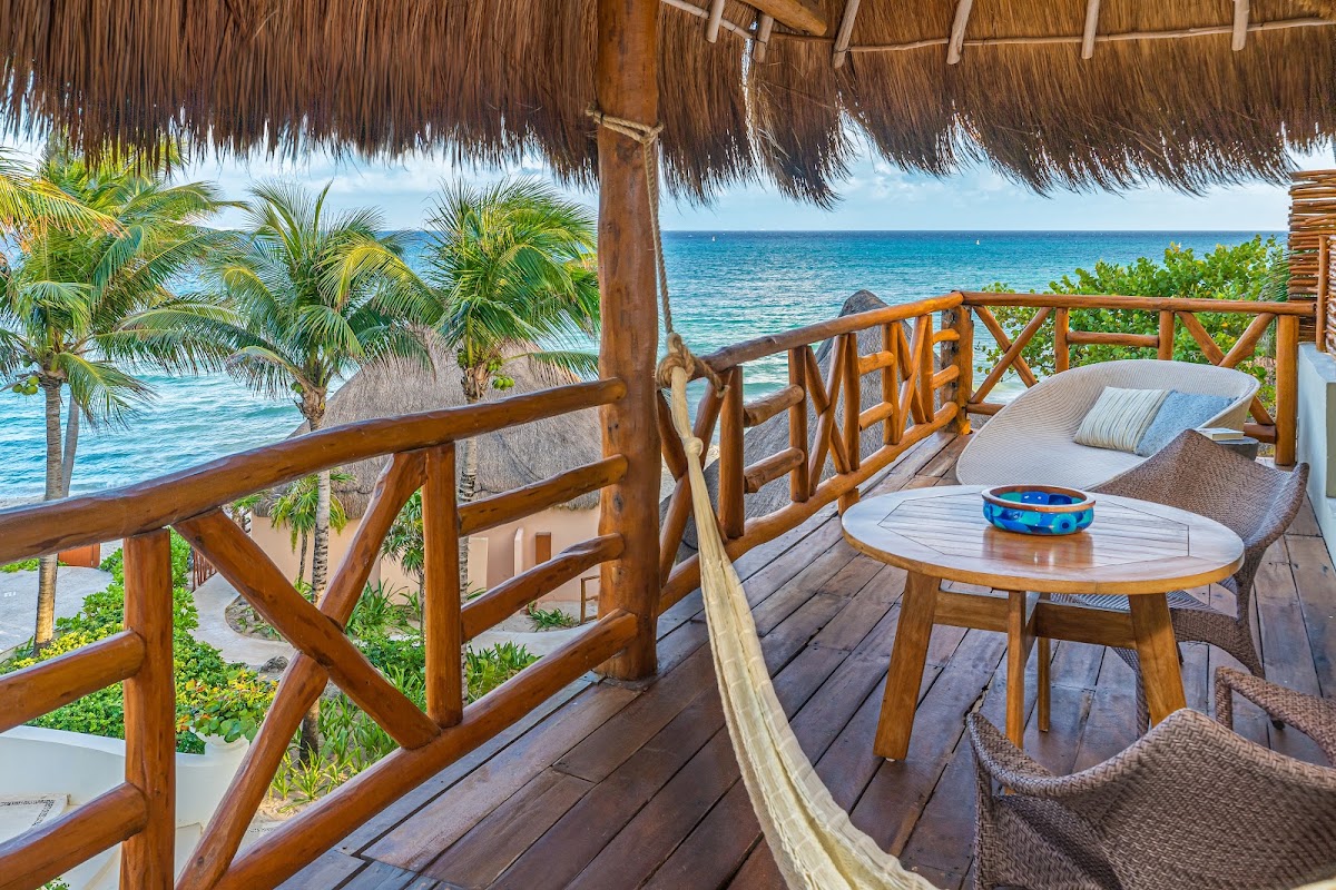 Wooden deck with thatched roof shelter overlooking turquoise ocean, palm trees, lounge chairs, and round table with blue bowl at Mahekal Beach Resort.