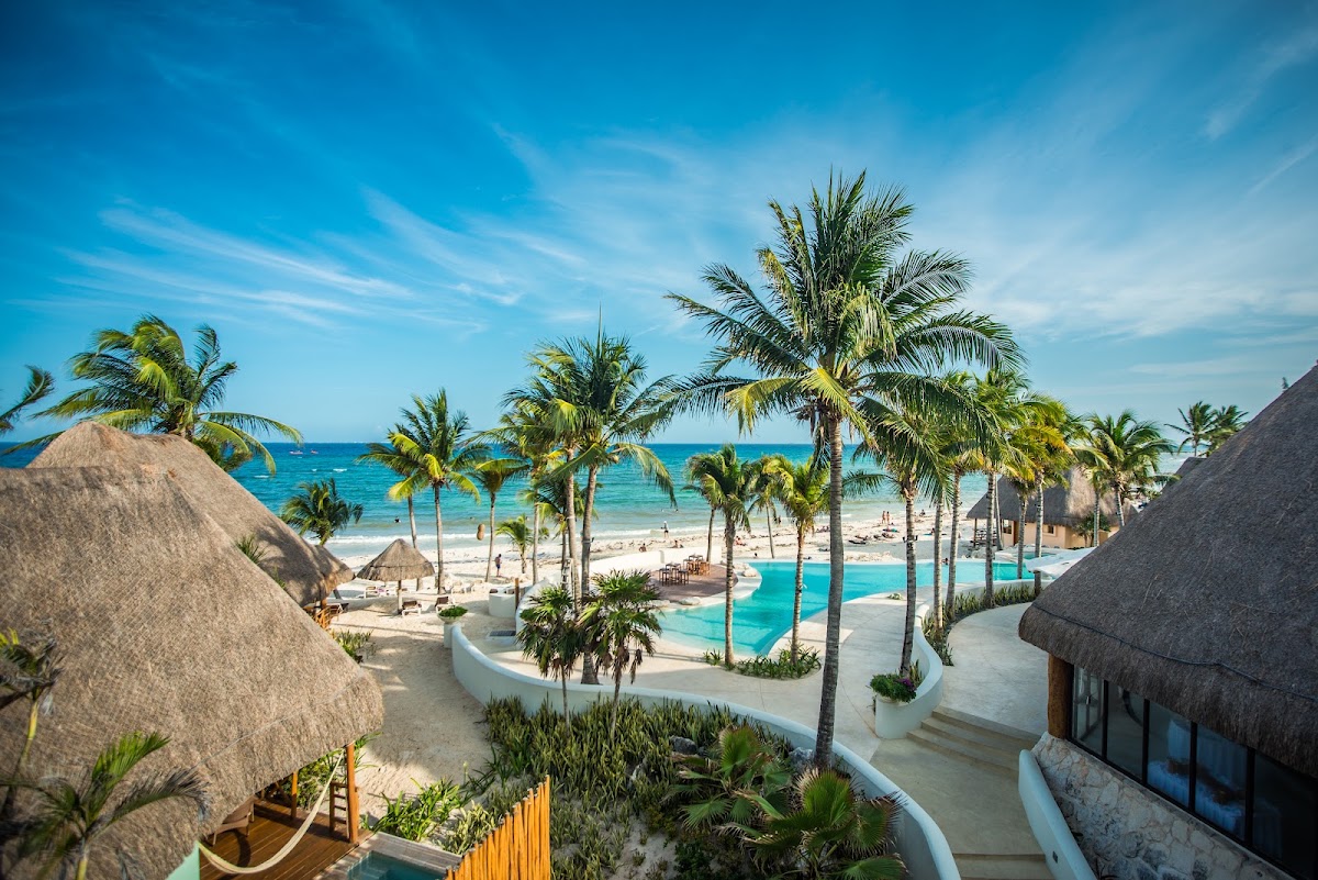 Thatched-roof bungalows frame a turquoise pool overlooking a sandy beach with palm trees and the blue ocean beyond at Mahekal Beach Resort.