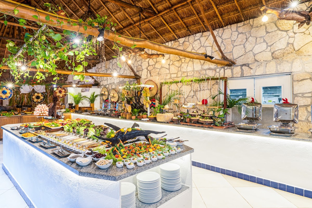 Mahekal Beach Resort's dining area features a palapa roof, stone walls, and buffet stations laden with salads, seafood, and appetizers under warm lighting.