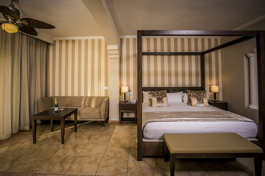 Spacious resort bedroom with black four-poster bed, cream striped walls, seating area, and ceiling fan.